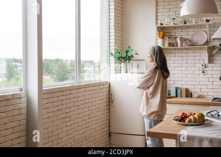 Image d'une jolie femme mûre debout à l'intérieur de la cuisine à la maison tout en regardant la fenêtre Banque D'Images