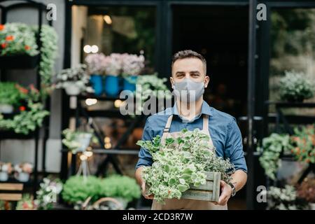 Petite entreprise et début de journée de travail. L'homme dans le masque de protection sort une boîte de plantes à l'extérieur Banque D'Images