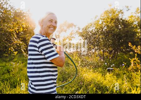 Heureuse femme âgée ayant plaisir arroser des plantes avec le tuyau dans le jardin d'été. Gouttes d'eau dans le rétroéclairage. Agriculture, jardinage, agriculture, vieux Banque D'Images