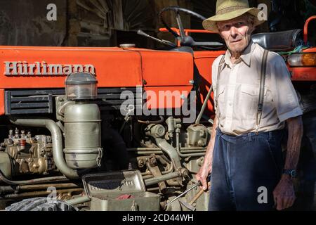 Un vieil agriculteur travaillant sur un tracteur rouge sous le toit de la maison de ferme au soleil. Vintage, vue latérale. Banque D'Images
