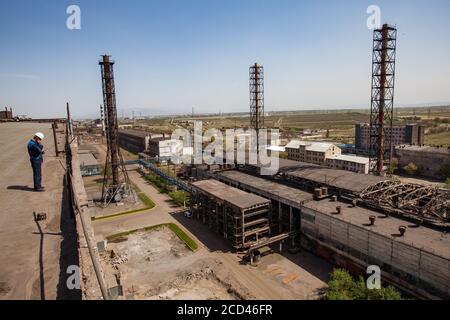 Anciens bâtiments non utilisés de l'usine de métallurgie soviétique et cheminées rouillées sur le ciel bleu. Ouvrier en casque blanc (à gauche) sur le toit. Ville de Taraz. Banque D'Images