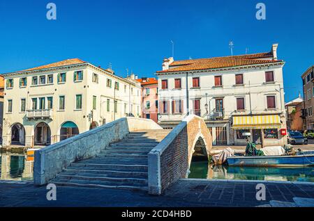 Pont en brique de pierre Ponte Pescheria à travers le canal d'eau de Vena et de vieux bâtiments dans le centre historique de la ville de Chioggia, ciel bleu en arrière-plan en été, région de Vénétie, Italie du Nord Banque D'Images