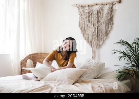 Jeune belle femme étudiante dans un t-shirt orange et jeans bleu assis sur le lit avec des oreillers et écoute un livre audio, musique, podcast sur le casque. Banque D'Images
