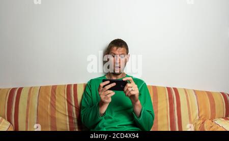 Le jeune homme est choqué de regarder le téléphone mobile sur le canapé. Portrait surprisé et étonné Banque D'Images