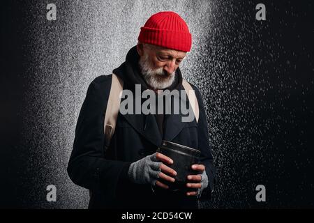 Vieux homme portant des vêtements noirs de rue et tenant une tasse de fer pour l'argent, pour l'aide. Triste homme sans abri sous forte pluie. Banque D'Images