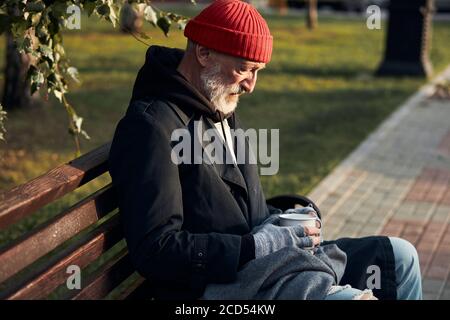 Homme à barbe grise sans abri assis dans des vêtements de rue sur le banc, sans nourriture et sans argent. Un homme qui courait a baissé la tête. Vue latérale sur les sans-abri Banque D'Images