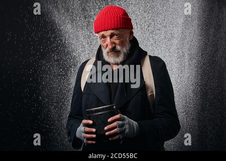 Vieux homme portant des vêtements noirs de rue et tenant une tasse de fer pour l'argent, pour l'aide. Triste homme sans abri sous forte pluie. Banque D'Images