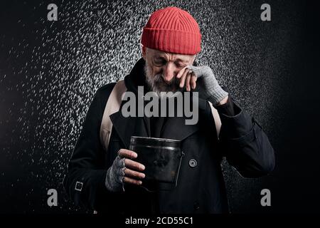 Vieux barbu portant des vêtements de rue noirs et tenant une tasse de fer pour l'argent, pour l'aide. Triste homme sans abri sous forte pluie. Banque D'Images