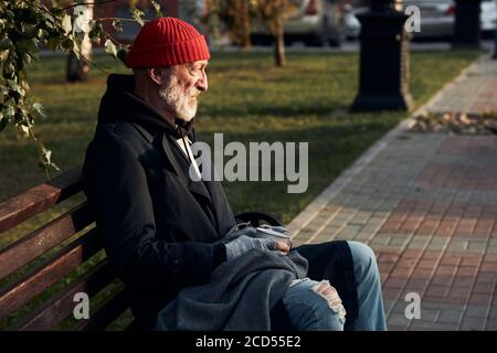 Homme mature sans abri assis dans des vêtements de rue sur le banc, sans nourriture et sans argent. Un homme qui courait a baissé la tête. Vue latérale sur le perso sans-abri Banque D'Images