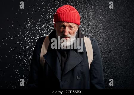 Vieux barbu portant des vêtements de rue noirs et tenant une tasse de fer pour l'argent, pour l'aide. Triste homme sans abri sous forte pluie. Banque D'Images