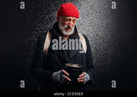 Vieux homme portant des vêtements noirs de rue et tenant une tasse de fer pour l'argent, pour l'aide. Triste homme sans abri sous forte pluie. Banque D'Images