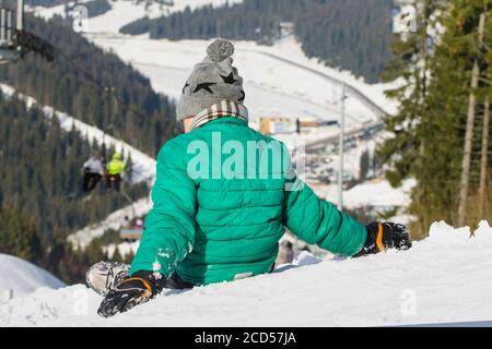 Garçon est assis sur une colline enneigée sur fond de téléphérique, de forêt de pins et de montagnes. Station de ski. Jour d'hiver Banque D'Images