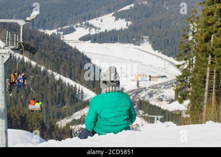 Garçon est assis sur une colline enneigée sur fond de téléphérique, de forêt de pins et de montagnes. Station de ski. Jour d'hiver. Banque D'Images