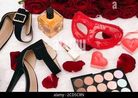 Masque de carnaval rouge, bouquet de roses rouges, chaussures noires avec talons, bougies en forme de coeur, rouge à lèvres, bouteille de parfum, ombre à paupières et éparpillées Banque D'Images