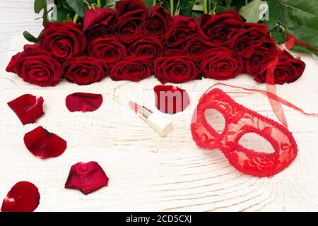 Concept de carnaval romantique. Masque de carnaval rouge, bouquet de roses rouges et rouge à lèvres sur fond de bois clair Banque D'Images