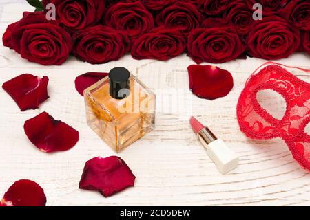 Concept de carnaval romantique. Masque de carnaval rouge, bouquet de roses rouges, rouge à lèvres et bouteille de parfum sur fond de bois clair Banque D'Images