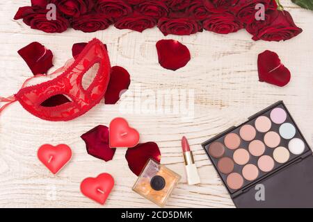 Concept de carnaval romantique. Masque de carnaval rouge, bouquet de roses rouges, rouge à lèvres, bouteille de parfum et ombre à paupières sur fond de bois clair Banque D'Images