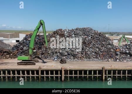 Une grande pile de ferraille attend l'expédition sur une jetée côté quai. Les machines industrielles sont visibles et utilisées pour trier les matériaux. Banque D'Images