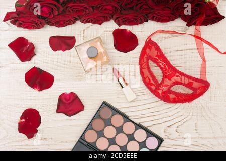 Concept de carnaval romantique. Masque de carnaval rouge, bouquet de roses rouges, rouge à lèvres, bouteille de parfum et ombre à paupières sur fond de bois clair Banque D'Images