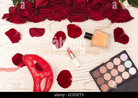 Concept de carnaval romantique. Masque de carnaval rouge, bouquet de roses rouges, rouge à lèvres, bouteille de parfum et ombre à paupières sur fond de bois clair Banque D'Images