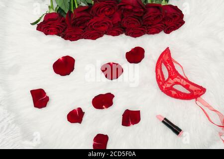 Concept de carnaval romantique. Masque de carnaval rouge, bouquet de roses rouges, rouge à lèvres et pétales épars sur la fourrure blanche. Banque D'Images