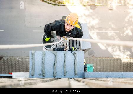 Un travailleur mâle suspendu à un projet d'accès à la corde et de construction en hauteur. Travail sur l'isolation des murs de construction et la peinture de façade décoration travail. Recherche Banque D'Images