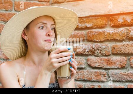 Jeune femme en grand chapeau de paille tenant un cocktail et fixe dans la surprise au verre. Fille avec boisson sur fond de mur de brique. Banque D'Images