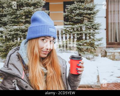 Une fille aux cheveux rouges dans un chapeau bleu tient un papier verre rouge avec couvercle en plastique noir sur fond de sapins et de chasse-neige Banque D'Images