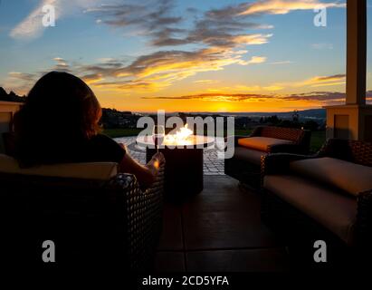 Une femme se détend avec un verre de vin la nuit en face d'un foyer extérieur sur un patio de une maison de luxe donnant sur une ville et une vallée Banque D'Images
