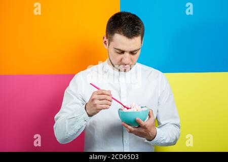 Un homme sur fond jaune, bleu, orange, rose dans une chemise blanche avec un plateau turquoise de guimauves et une cuillère rose. Le gars avec un sourire. Un im Banque D'Images