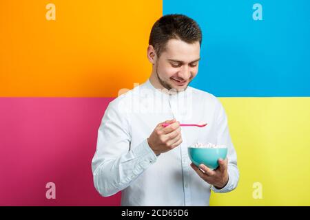 Un homme sur fond jaune, bleu, orange, rose dans une chemise blanche avec un plateau turquoise de guimauves et une cuillère rose. Le gars avec un sourire. Un im Banque D'Images