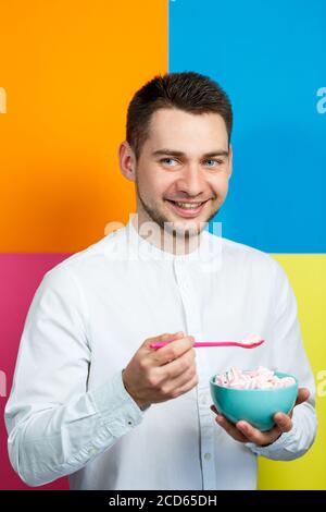Un homme sur fond jaune, bleu, orange, rose dans une chemise blanche avec un plateau turquoise de guimauves et une cuillère rose. Le gars avec un sourire. Un im Banque D'Images