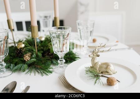 Table de Noël ou du nouvel an. Assiettes, coutellerie argentée, verres, boîte-cadeau, décoration de branche festive, bougies, cerf brillant sur un fond de table blanc Banque D'Images