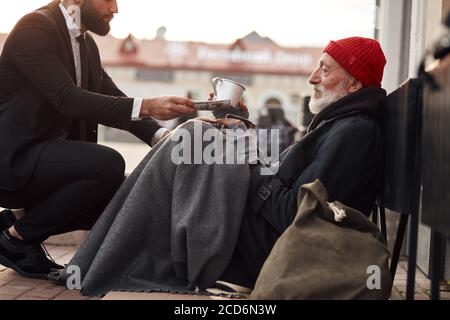 Un homme gentil en costume dédort aux sans-abri et donne de l'argent de don, une facture d'un dollar à mendiant mâle Banque D'Images
