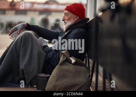 Vue latérale sur le mendiant mâle dans la rue mendiant, attendant l'aide des citoyens. Un homme sans domicile en vieux vêtements est assis sur le sol par temps froid Banque D'Images