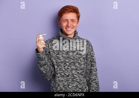 positif drôle gars avec un sourire en gris chaud chandail avec pulvérisation nasale. portrait de gros plan, arrière-plan bleu isolé, prise de vue en studio Banque D'Images