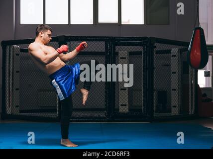 Jeune homme sportif se préparant pour les compétitions de kickboxing, se réchauffant dans la salle de gym en faisant des coups de pied avec les jambes pendant l'entraînement Banque D'Images