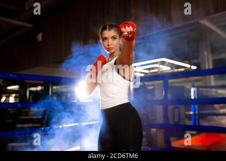 Jeune fille de mma caucasienne en uniforme de sport prêt à combattre en anneau de boxe, bandages rouges sur ses poings, préparation de combat de la femelle sportive musculaire Banque D'Images