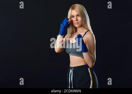 Magnifique boxer femme blonde déterminée dans des enveloppements de main bleus se préparer pour le grand combat, faire l'entraînement cardio-boxe dans vide sombre studio avec fumé Banque D'Images