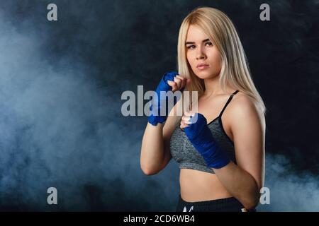 Magnifique boxer femme blonde déterminée dans des enveloppements de main bleus se préparer pour le grand combat, faire l'entraînement cardio-boxe dans vide sombre studio avec fumé Banque D'Images