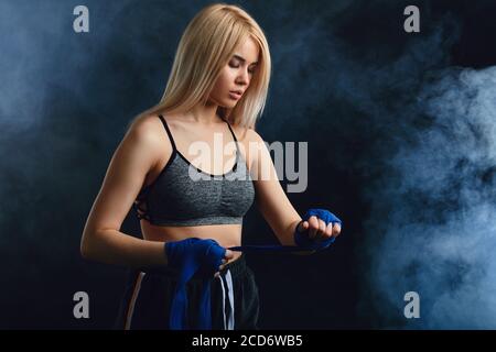 L'athlète blonde de sexe féminin s'exerce en défense et attaque sur un arrière-plan sombre et fumé. La femme dans les bandages de char-top et de boxe a le wor de boxe cardio Banque D'Images
