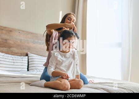 Ludique attrayant jeune maman volontiers peignes brunette cheveux de petite jolie fille assis sur le lit dans la chambre lumineuse, tons pastel, fond de vent vif Banque D'Images