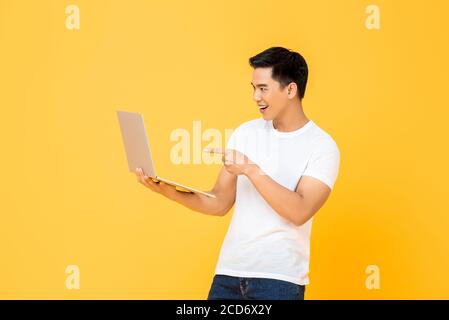 Portrait d'un jeune homme asiatique souriant tenant un ordinateur portable ordinateur tout en regardant et pointant sur l'écran dans un studio isolé arrière-plan jaune Banque D'Images