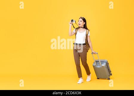 Portrait de voyage pleine longueur de jeune touriste enthousiaste asiatique femme marche tout en prenant des photos avec l'appareil photo dans un studio isolé jaune arrière-plan Banque D'Images