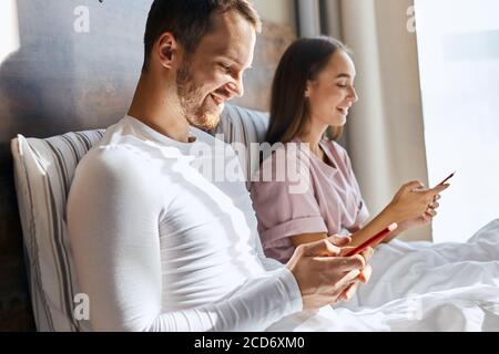 Une jeune femme et un mari agréables qui discutent avec leurs parents à l'aide d'un téléphone portable, qui reçoivent de bonnes nouvelles, qui sourient largement, qui regardent de côté avec des visages heureux, dans une chambre à coucher Banque D'Images