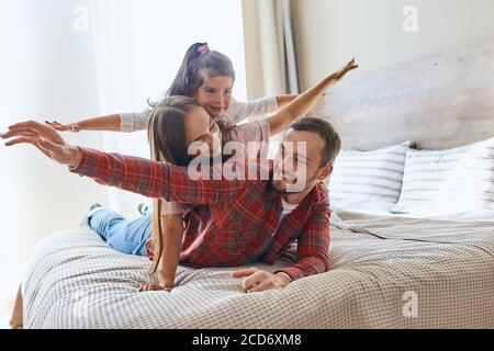 Famille heureuse et aimante passant du temps ensemble, père, mère et fille jouant gaiement sur le grand lit dans la chambre d'hôtel, étirant les mains, s'amuser, smi Banque D'Images