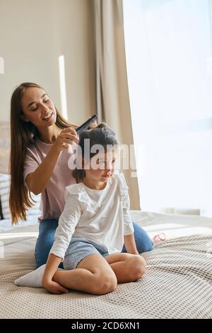 Petite fille brune vêtue de façon décontractée, assise sur les genoux tandis que la mère peignant ses cheveux, regardant loin, exprimant le calme, fatigué d'attendre pour la coiffure, Banque D'Images