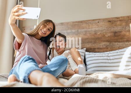 Famille heureuse et aimante. Mère et enfant fille jouant, embrassant, prenant des photos sur téléphone cellulaire, allongé sur le lit, regardant loin, concept Happy Morning Time, botto Banque D'Images