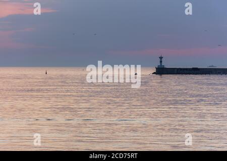 Un phare contre le ciel de coucher de soleil. Magnifique paysage marin paisible. Petits navires à l'horizon. Nuages violets et eau douce rose. Port maritime. Goélands dedans Banque D'Images