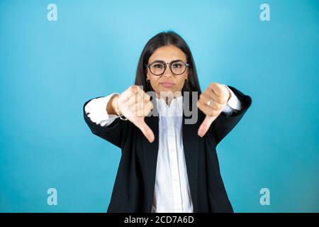 Jeune belle femme d'affaires sur un fond bleu isolé avec le visage en colère, signe négatif montrant déplaît avec les pouces vers le bas Banque D'Images